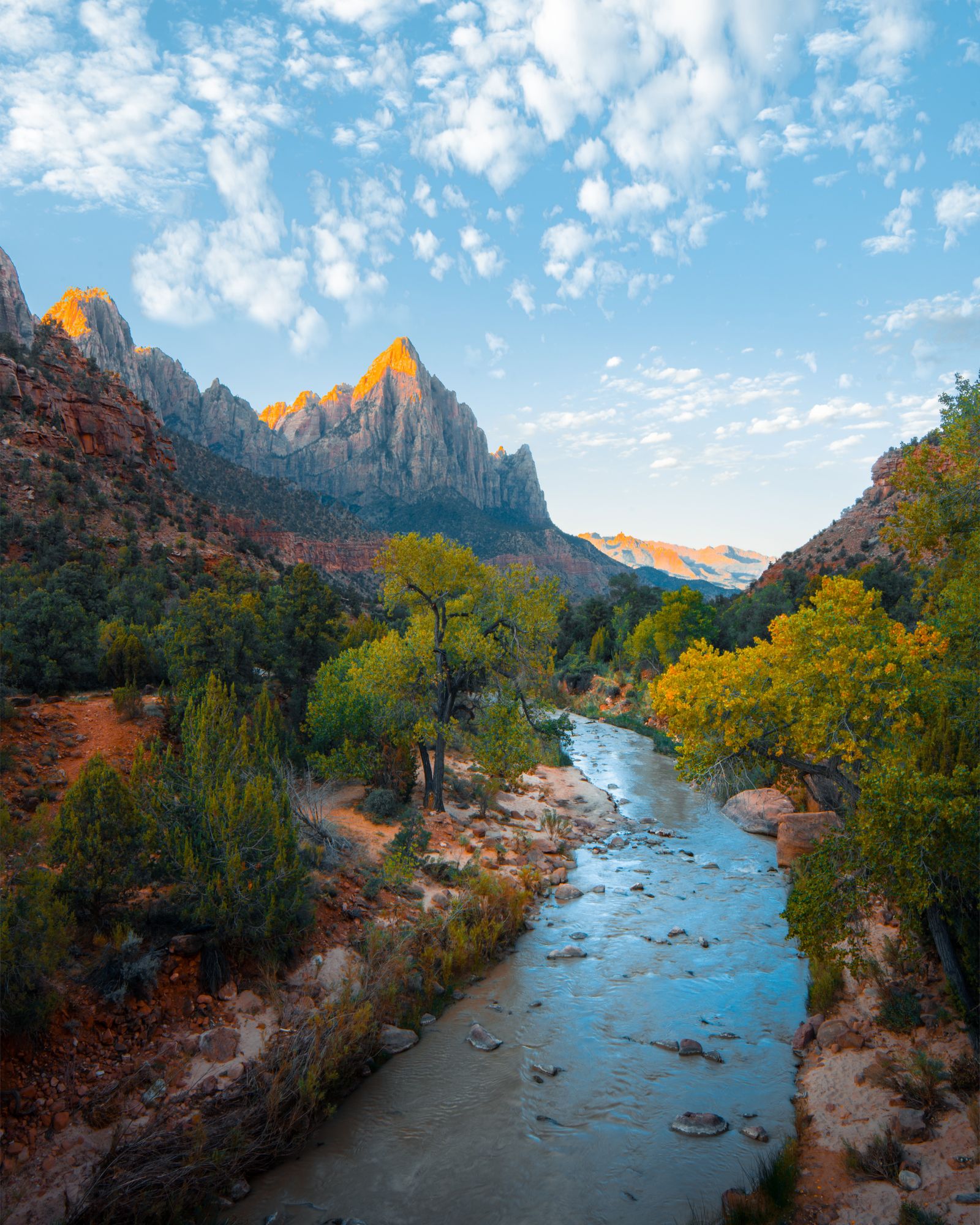 River winding through a canyon with yellow autumn trees and a sunlit mountain peak under a cloudy blue sky