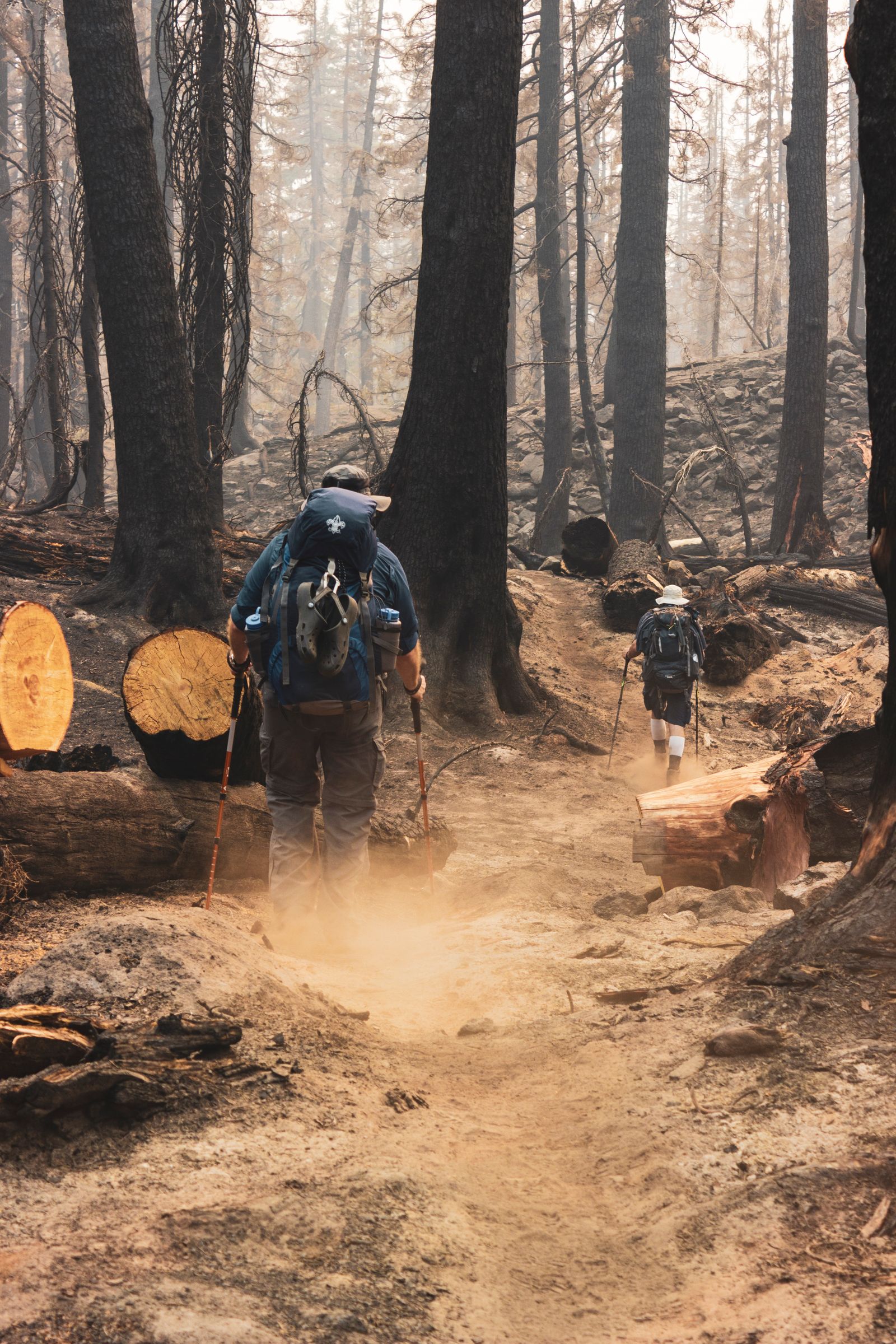 Two hikers with backpacks and orange trekking poles on a dusty trail through charred forest trees